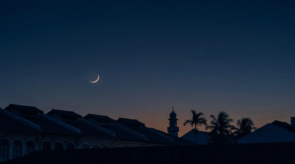 A thin new crescent moon in a deep navy twilight sky above the silhouetted rooflines of tropical shophouses, a minaret and palm trees.
