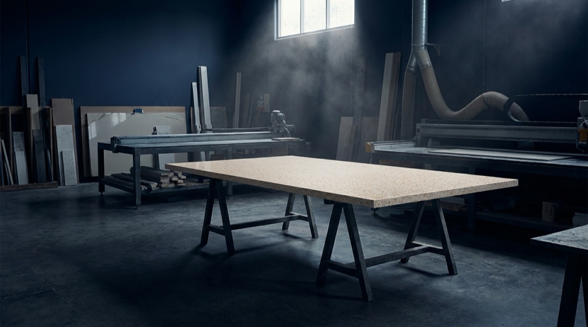 A manufactured composite slab raised on trestles in a stone fabrication workshop, fine dust hanging in a shaft of window light