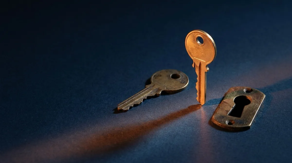 Photographic still life on a deep navy ground: a tarnished old key lying flat beside an upright new key catching sidelight, with a brass keyhole plate alongside