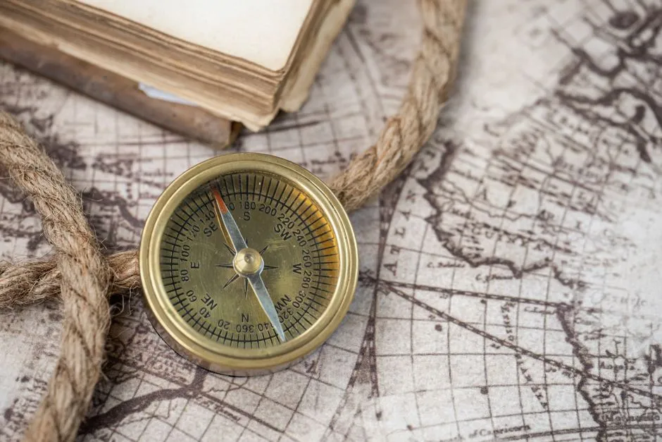 An antique brass compass with degree gradations and cardinal markings resting on an old map beside a rope and book