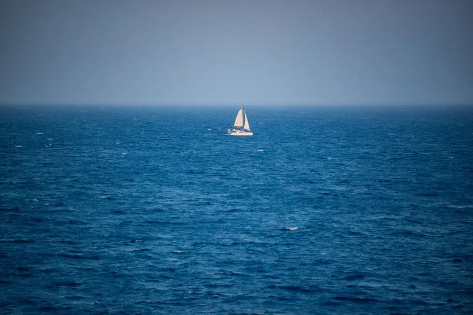 A lone white sailboat drifting on a wide open blue sea under a hazy sky
