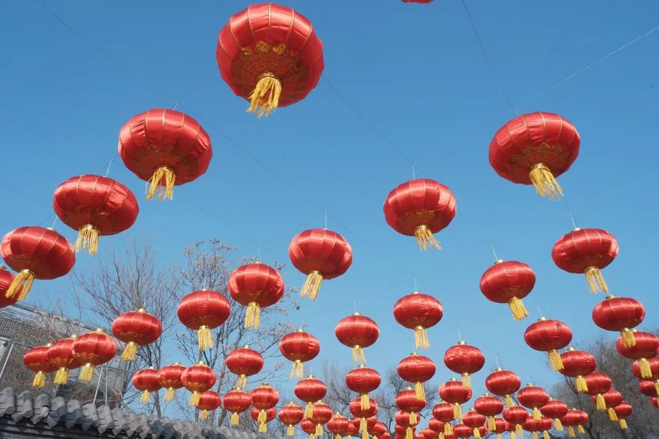 Rows of red Chinese lanterns with gold tassels strung against a clear blue sky above a tiled roof