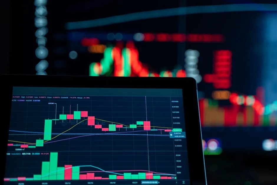 A tablet showing a red-and-green candlestick trading chart, with market screens glowing behind it