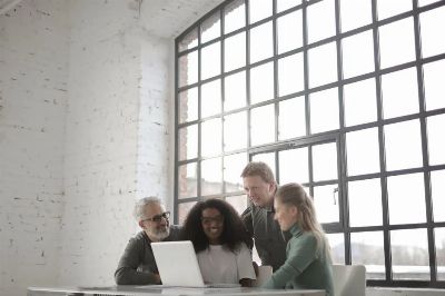 Positive focused multiracial coworkers gathering together near table with laptop in workplace at industrial building during remote work on team against big window at daytime