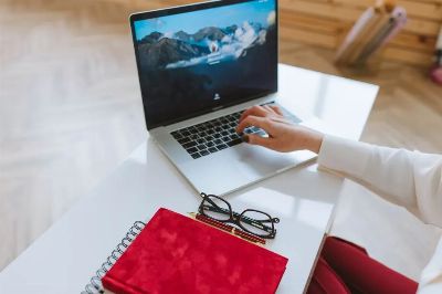 A person working on a laptop with a red notebook and glasses on a white table.