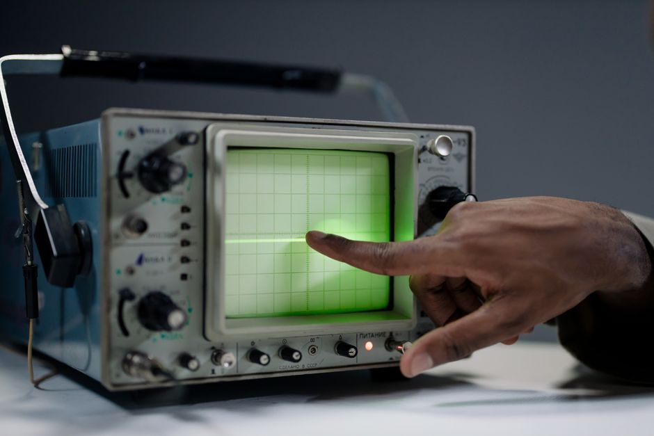 A hand points at the glowing green trace on the screen of a vintage oscilloscope in a dim room.