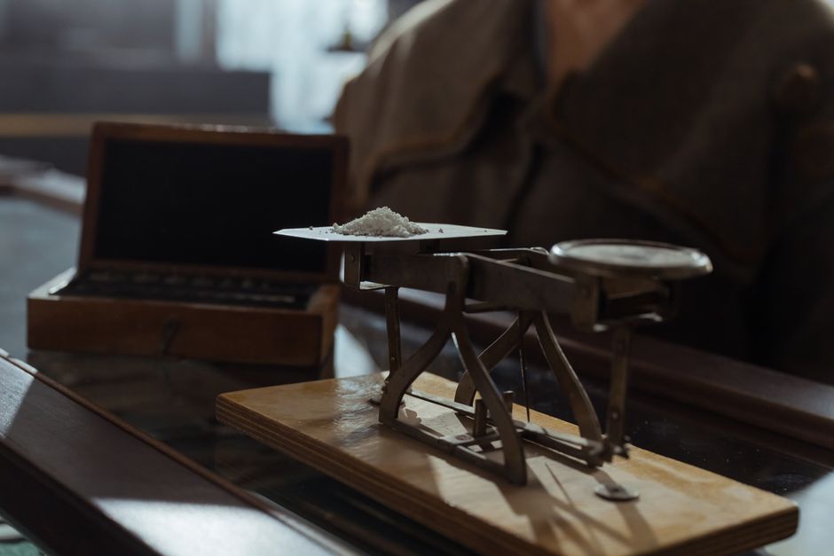A small antique beam balance on a wooden board holds a heap of white powder on one pan, lit low against a dark interior.