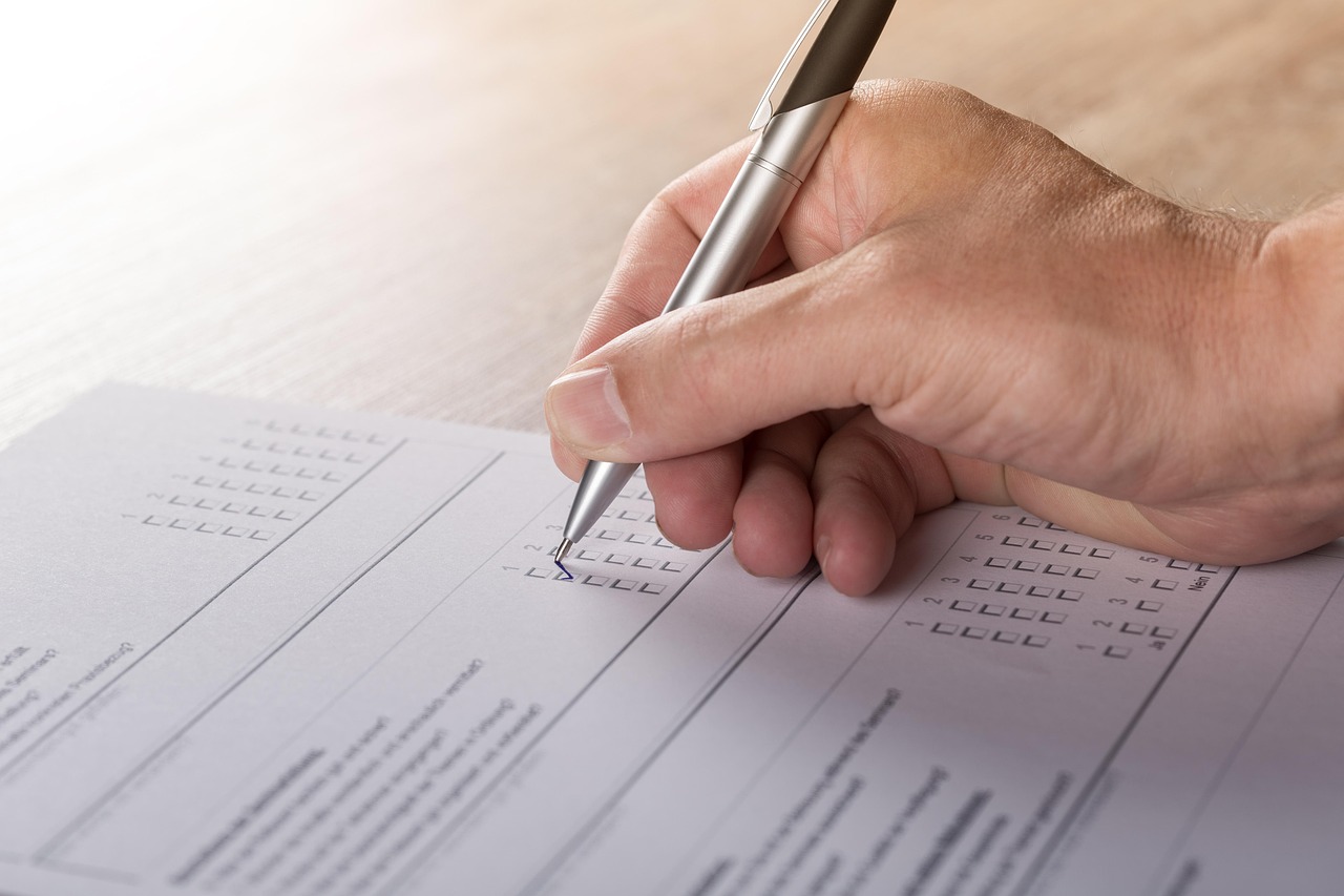 A hand holding a slim pen ticks boxes on a printed multiple-choice questionnaire on a pale desk.