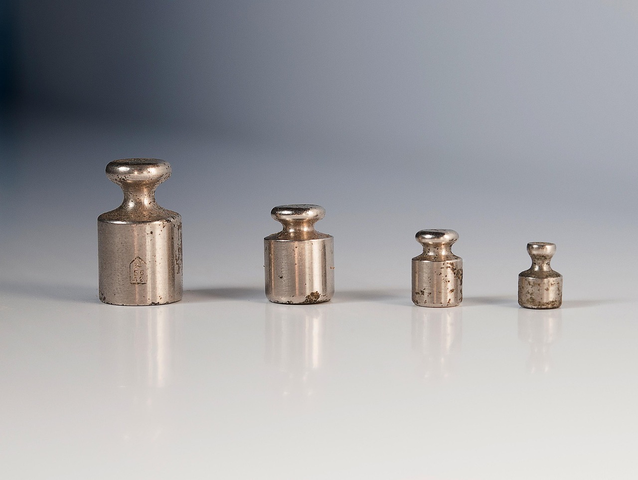 Four brass calibration weights of decreasing size stand in a row on a pale reflective surface.