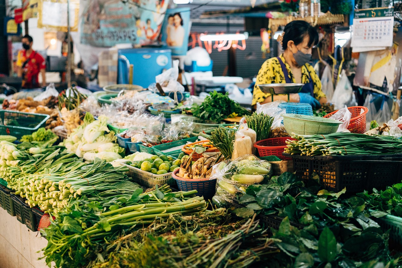 A busy wet-market stall piled with leafy greens, chillies and prepared foods, the vendor working behind it.