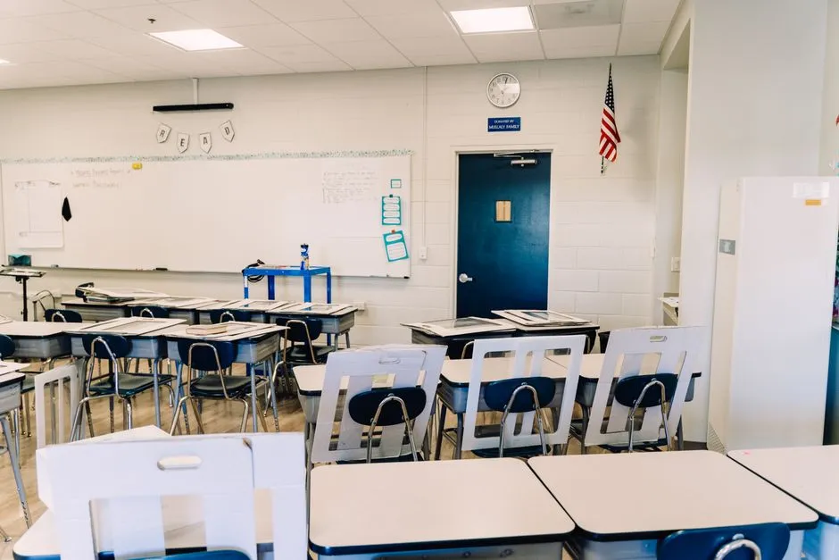 An empty American public school classroom with rows of single desks pushed together, a blank whiteboard, a wall clock and a United States flag by the door
