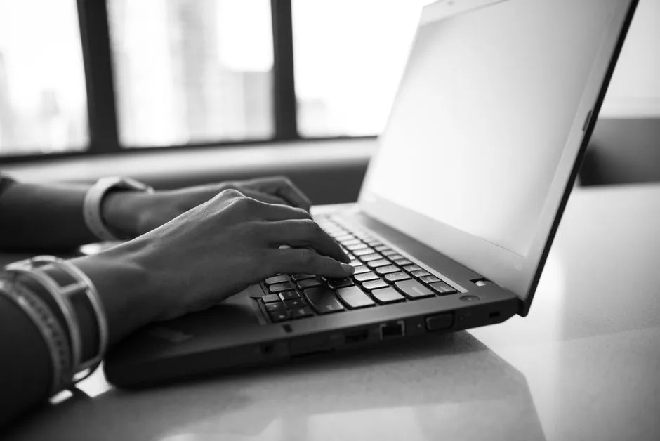 A black and white photograph of two hands typing on a laptop keyboard beside a bright window
