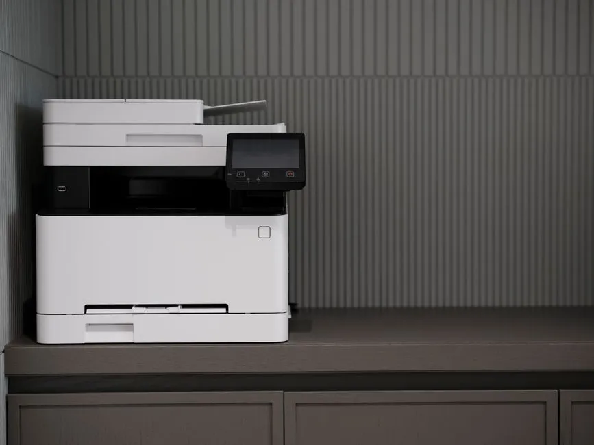 A white multifunction office printer with a small touchscreen, standing on a dark wooden cabinet.
