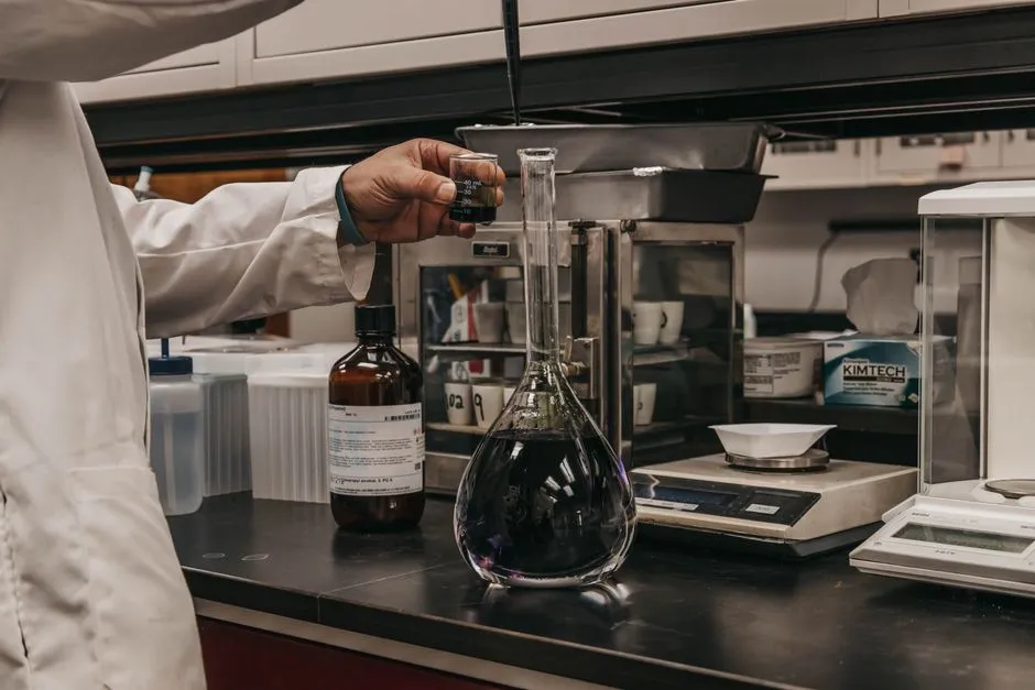 A gloved hand in a lab coat filling a volumetric flask of dark purple solution on a laboratory bench, an analytical balance beside it.