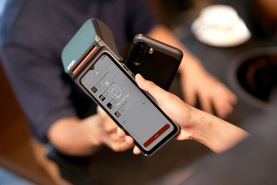A phone held against a card terminal to pay, seen over the shoulder of the cashier — illustrative of the tap-to-pay method the new balance uses.