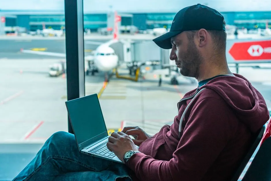 A traveller sitting at an airport gate with a laptop, an aircraft visible through the window behind.