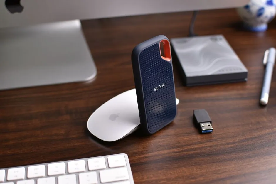 A portable solid-state drive and a small USB stick on a wooden desk beside a keyboard.