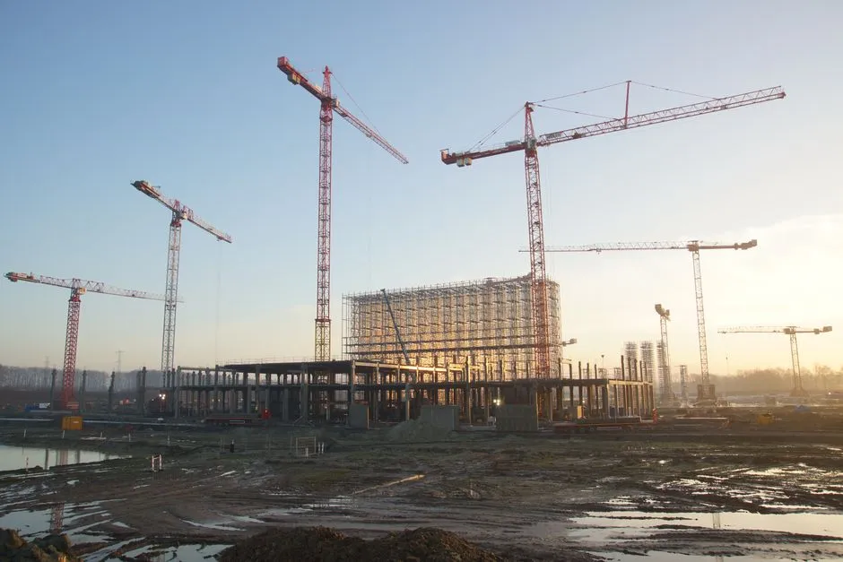 Tower cranes around a steel-framed building under construction on a large site, illustrating a report on data centre capital expenditure passing one trillion dollars.