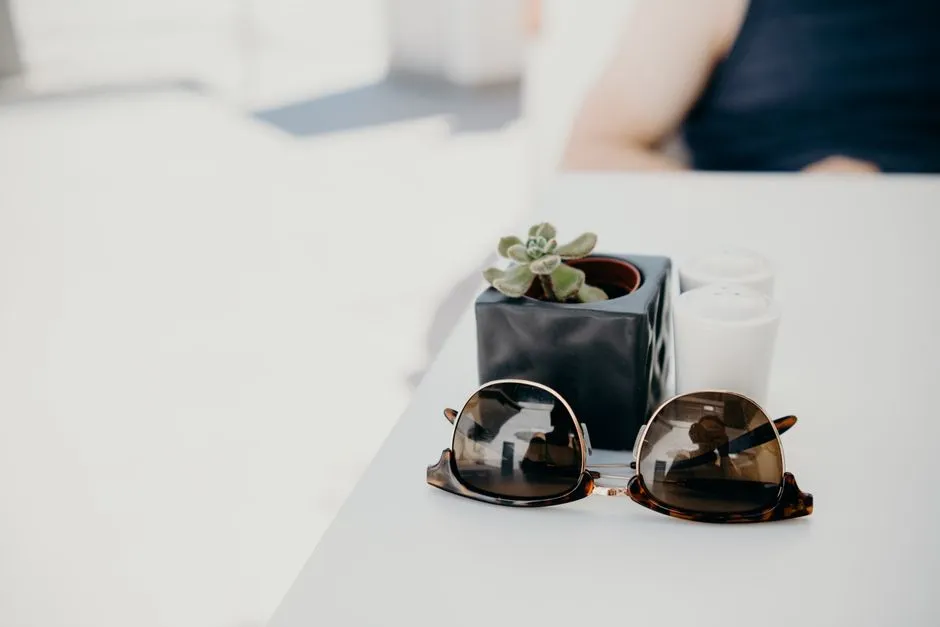 Two pairs of folded tortoiseshell sunglasses resting on a white table beside a small potted succulent, with a person seated out of focus behind them