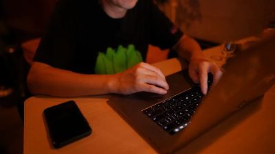 An adult man working at a desk on a laptop with a smartphone nearby, in a dimly lit home office.