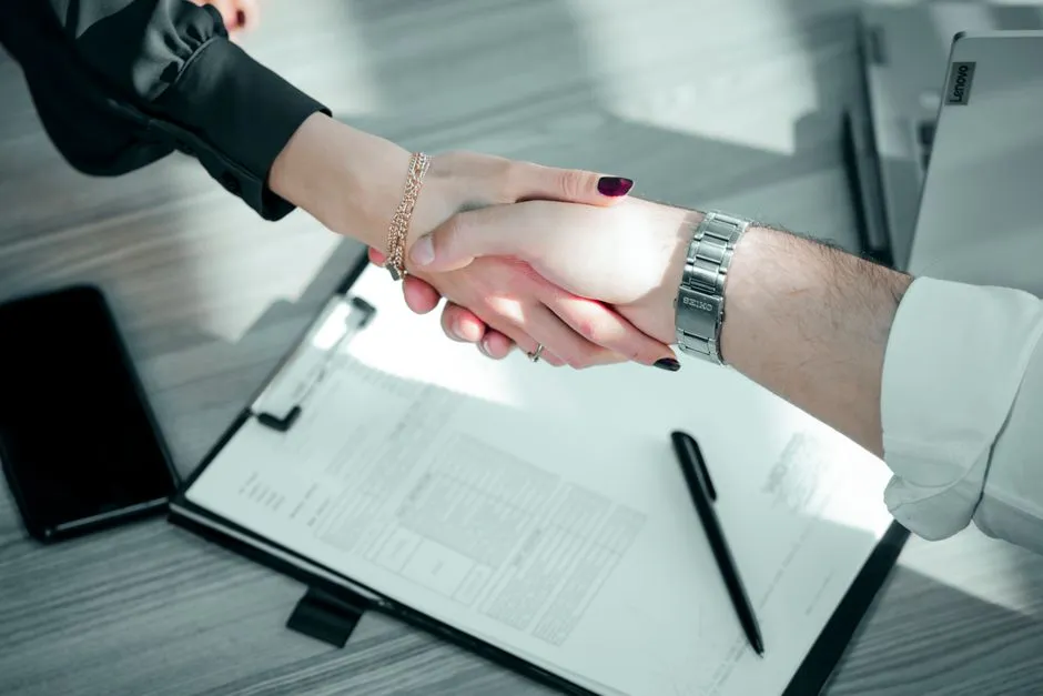 Two people shaking hands across a desk over an unsigned document on a clipboard, a pen lying beside it