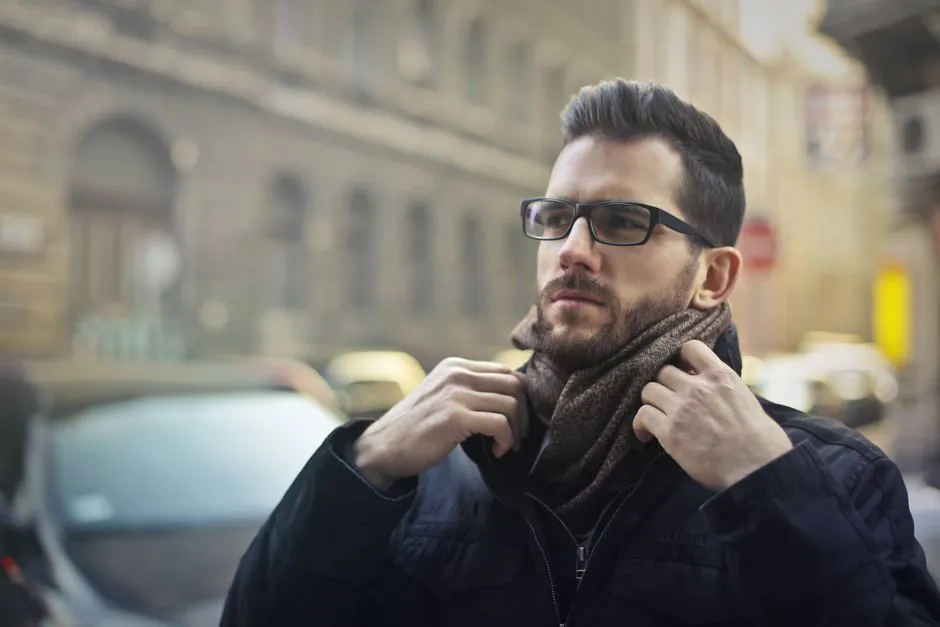 A man in ordinary spectacles adjusting his scarf on a city street, with traffic and buildings behind him