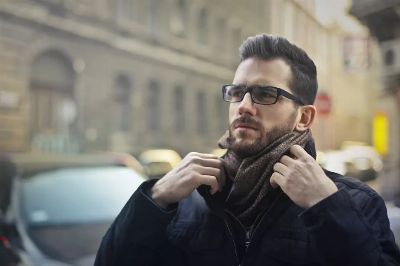 A fashionable man adjusting his scarf on a city street, wearing glasses and a dark jacket.