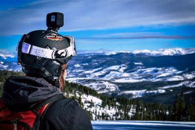 Person with helmet and camera overlooking snow-capped mountains and trees, capturing winter's beauty.