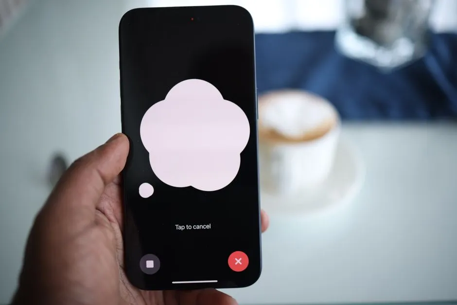 A hand holding a smartphone displaying a voice assistant listening screen, with a coffee cup blurred in the background