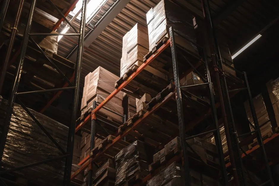 Tall warehouse racking stacked with palletised boxes, lit from above in a dim aisle