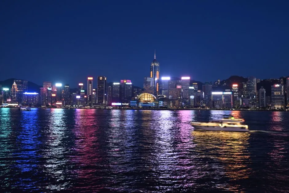 The lit towers of Hong Kong Island reflected across Victoria Harbour after dark, with a small ferry crossing the water in the foreground