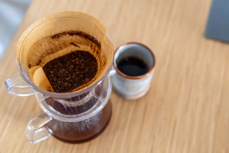 A glass pour-over coffee brewer on a wooden table, its paper cone still holding a bed of wet spent grounds while the brewed coffee sits dark in the carafe below, beside a small ceramic cup.