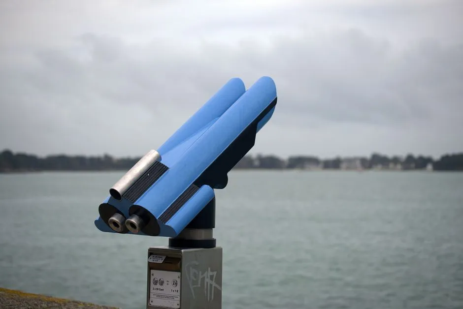 A blue coin-operated seafront viewer on a concrete plinth, pointed out across flat grey water under a heavy overcast sky, with a low treeline on the far shore.