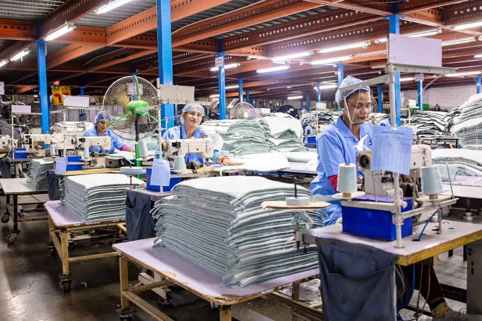 Workers in hairnets at sewing machines on a garment factory floor, with stacks of folded fabric on trolleys