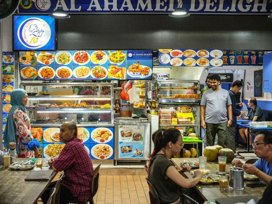 Diners at tables in front of a brightly lit food stall with a menu board of dishes