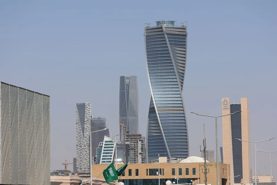A modern Gulf city skyline of glass towers under a pale hazy sky, including a tall twisting tower, with a green flag flying above low buildings in the foreground.