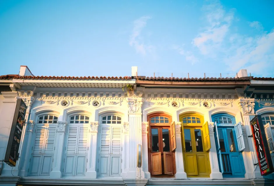 A row of Singapore shophouses photographed from the street — architecturally identical units under one continuous roofline, each painted a different colour: white, then red, yellow and blue.
