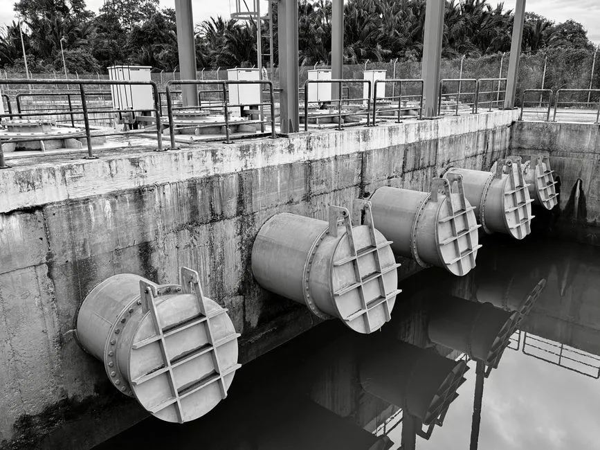 A row of large outfall pipes with sluice gates set into a concrete wall above still water, photographed in monochrome