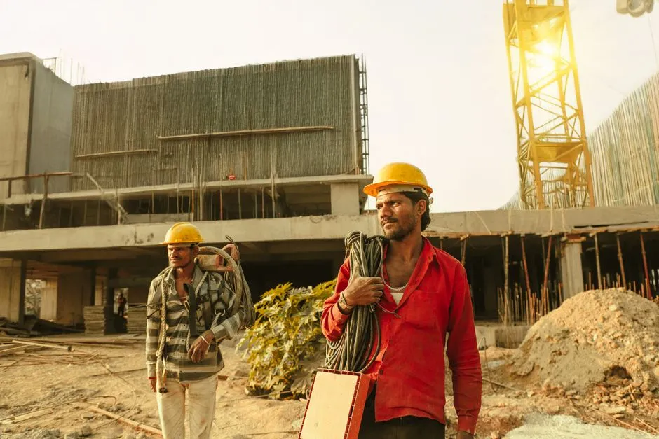 Construction workers in hard hats walking across a large building site, with a tower crane and exposed reinforcement bars behind them.