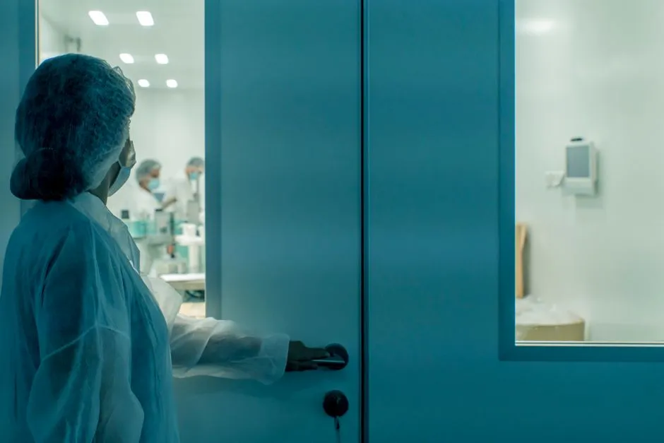 A person in a gown and hairnet standing at a sealed laboratory door, with colleagues visible through the glass panel beyond.