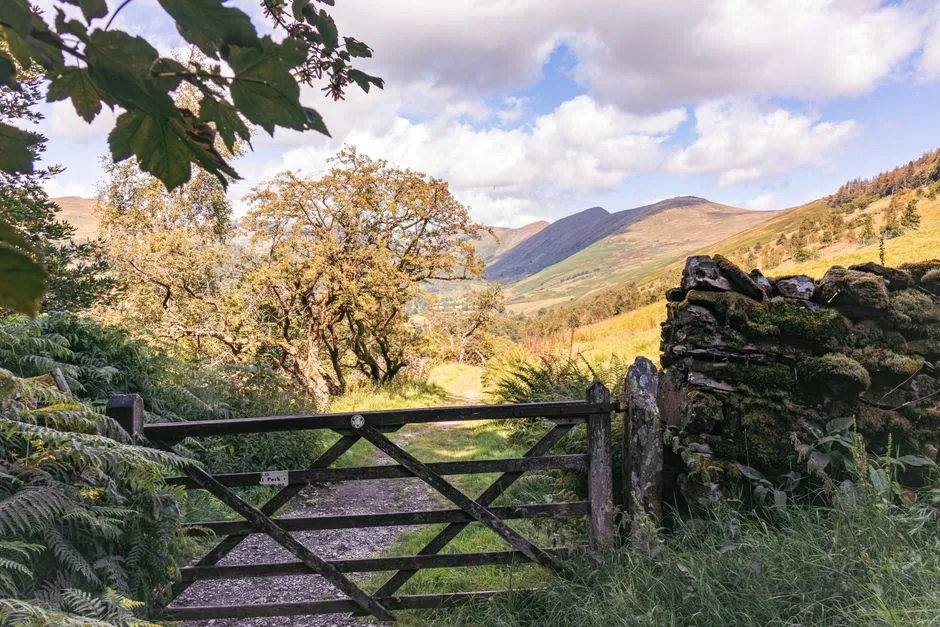 A closed wooden five-bar farm gate across a stone track, with open hills and a bright valley visible beyond it
