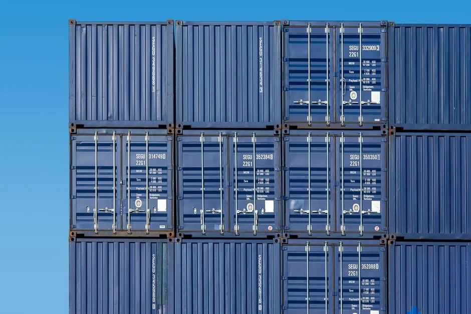 Identical blue shipping containers stacked in a grid against a clear sky — illustrative of a standard packaging format, where a uniform outer shape is what lets the contents move between systems.
