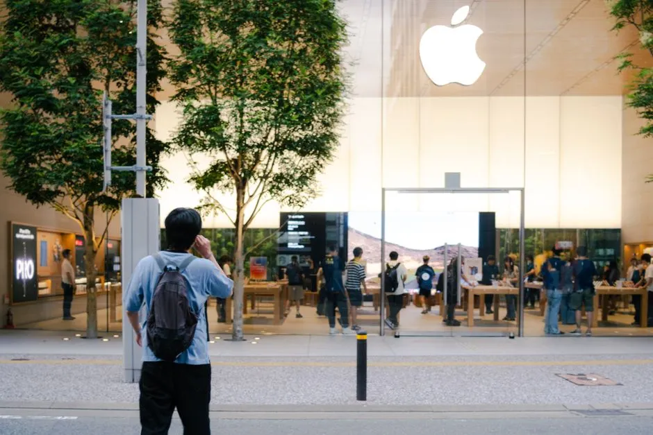 A busy Apple Store entrance with glass facade, trees, and people milling around.