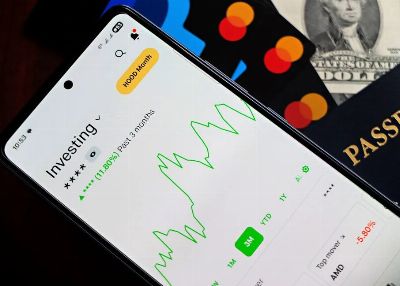 A smartphone showing an investment app with green growth indicators, surrounded by credit cards, US dollars, and a passport.