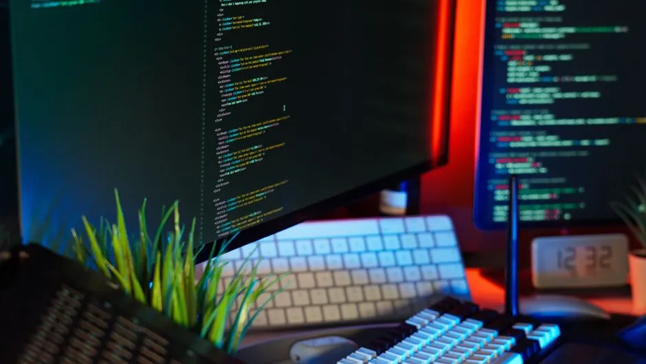 A developer's desk at night: two monitors filled with coloured code, two keyboards and a small desk clock reading 12:32, lit red and blue