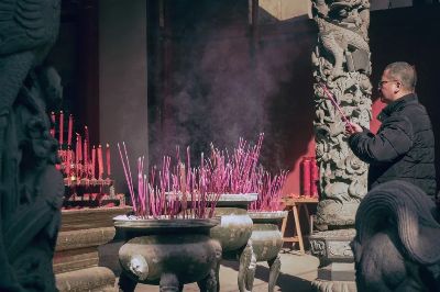A man lights incense sticks in an Asian temple, surrounded by intricate sculptures.