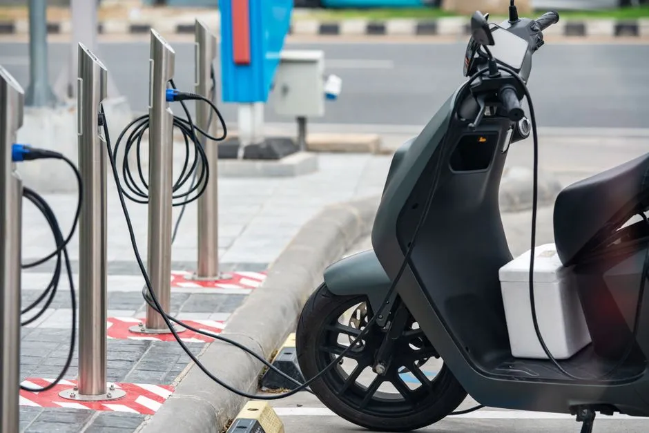An electric scooter plugged into a row of kerbside charging posts, its cable running across the pavement