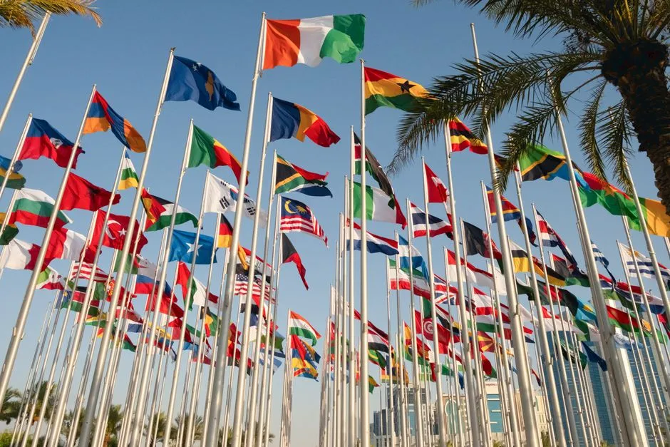 National flags of many countries flying on poles against a clear blue sky.