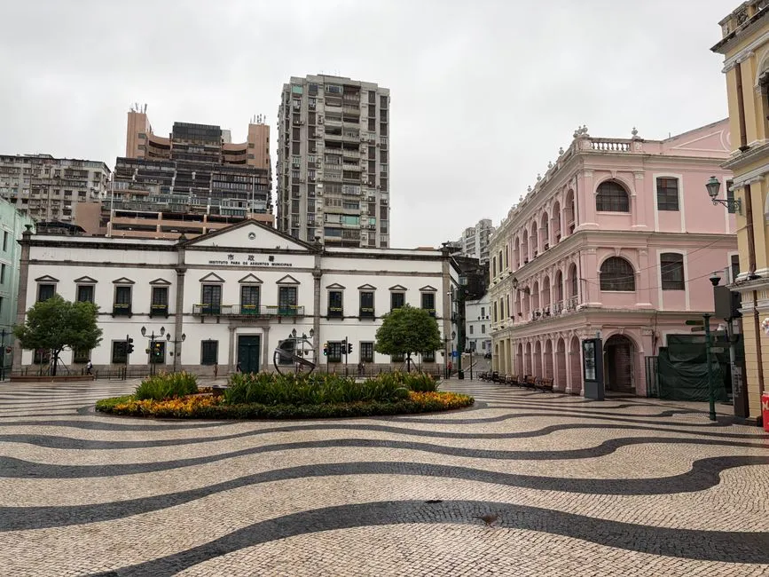 The wave-patterned Portuguese paving of Senado Square in Macau, with the white Leal Senado building on the left and a pink colonial-era building on the right under an overcast sky.