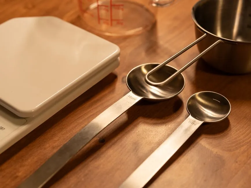 Stainless steel measuring spoons marked 15 ml and 5 ml lying on a wooden worktop beside a digital kitchen scale, a glass measuring jug and a metal measuring cup.